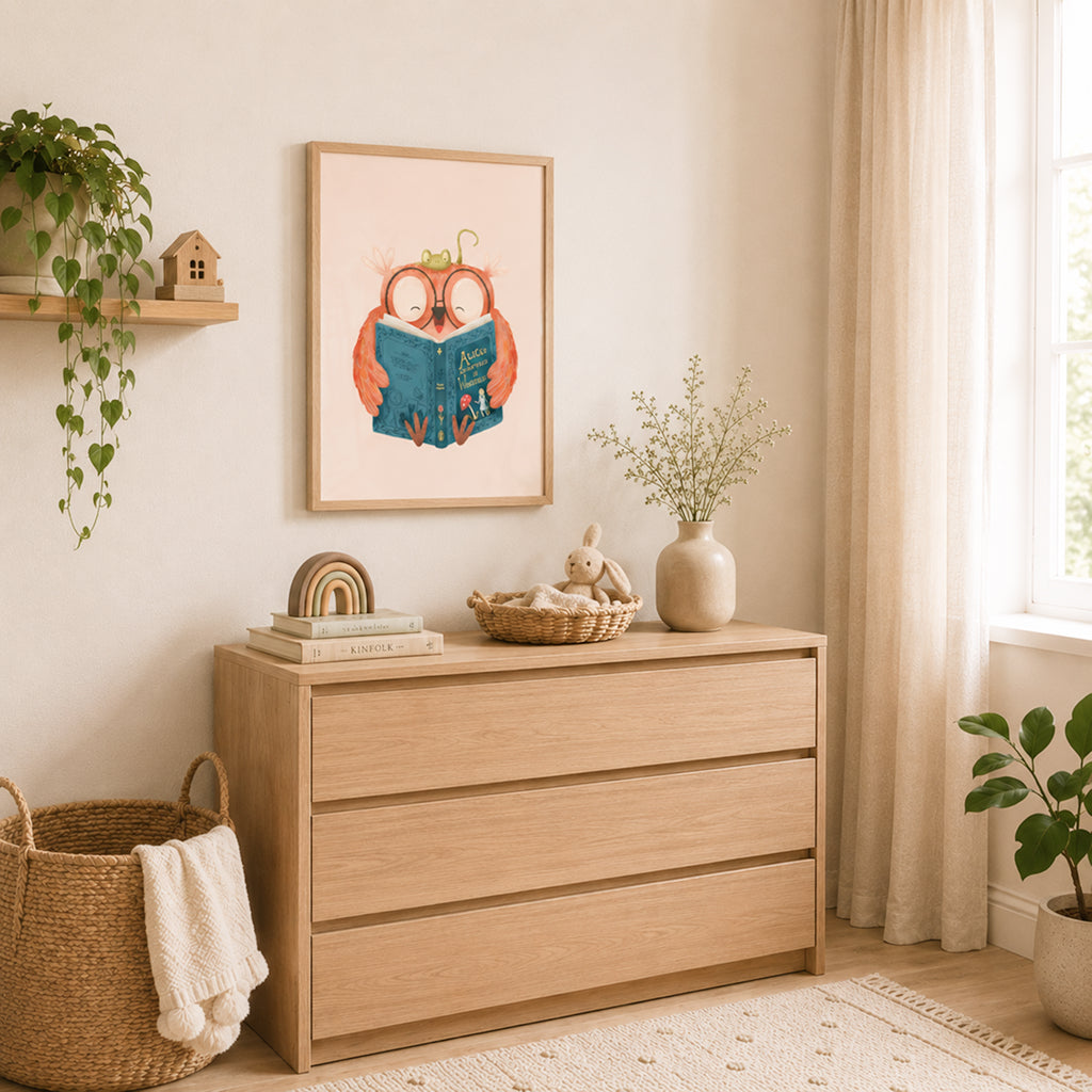 Cozy Scandinavian-style children's room with a light oak dresser beneath a framed owl illustration. The poster features a cheerful owl reading a blue storybook with a small green frog resting on its head against a soft blush-pink background. Natural light streams through sheer curtains, illuminating woven baskets, a rainbow stacking toy, a vase with delicate flowers, and green plants throughout the room.