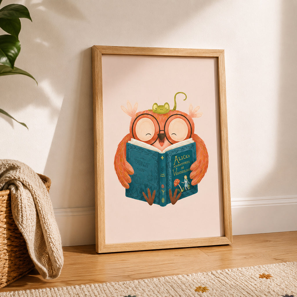 Framed owl wall art print in a cozy Scandinavian-style children's room. The illustration shows a cheerful owl reading a blue book, displayed in a light wood frame leaning against a wall, with soft natural sunlight, a woven basket, and neutral-toned decor.