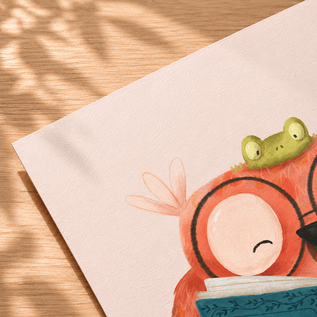 Close-up of a textured art print resting on a warm wooden surface with soft sunlight and plant shadows. The lower-right corner of the illustration shows part of a cheerful owl reading a blue book, with a small green frog perched on its head. The print features a soft blush-pink background and visible paper texture.